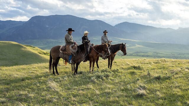 Three horseback riders crossing lush, rolling grassland on the 7,400-plus-acre Cato Ranch, with the Bighorn Mountain foothills in the background